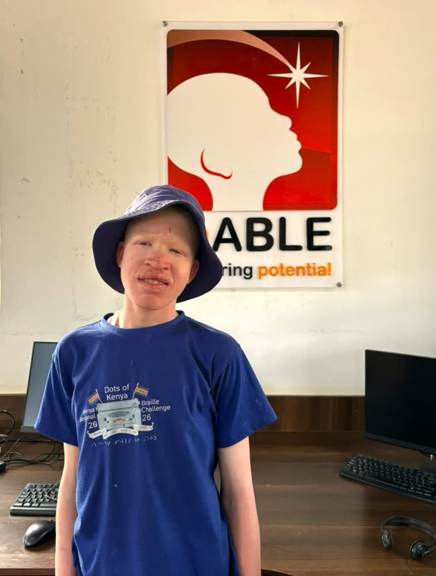Eric is wearing a blue sun hat and a Kenya National Braille Challenge T-shirt and stands in a computer lab with desktop computers and an inABLE sign in the background.