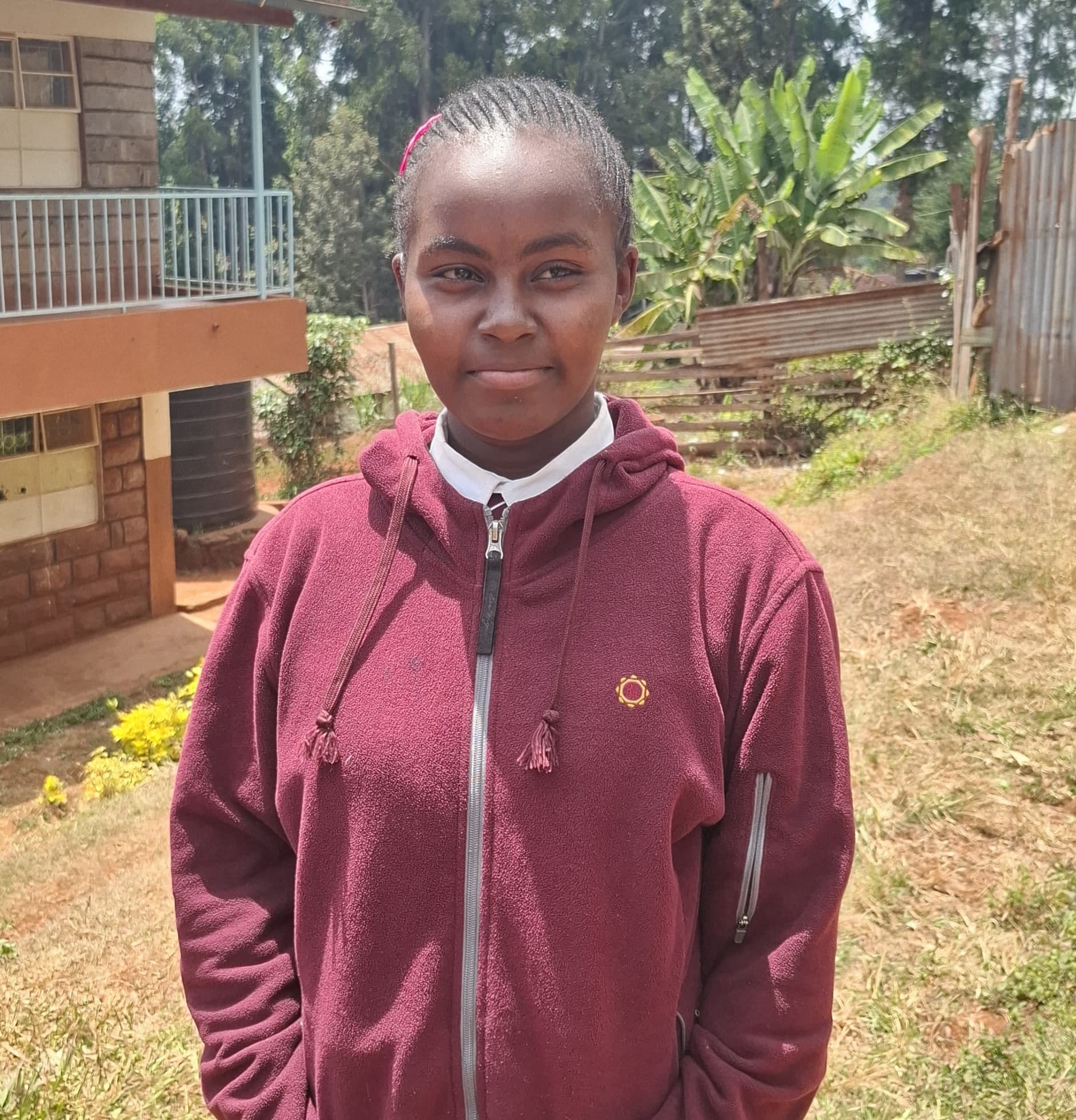 Sharin stands outdoors on a grassy school compound, wearing a maroon zip-up school sweater over a white collared shirt and a plaid skirt. The student is facing the camera with hands clasped in front.